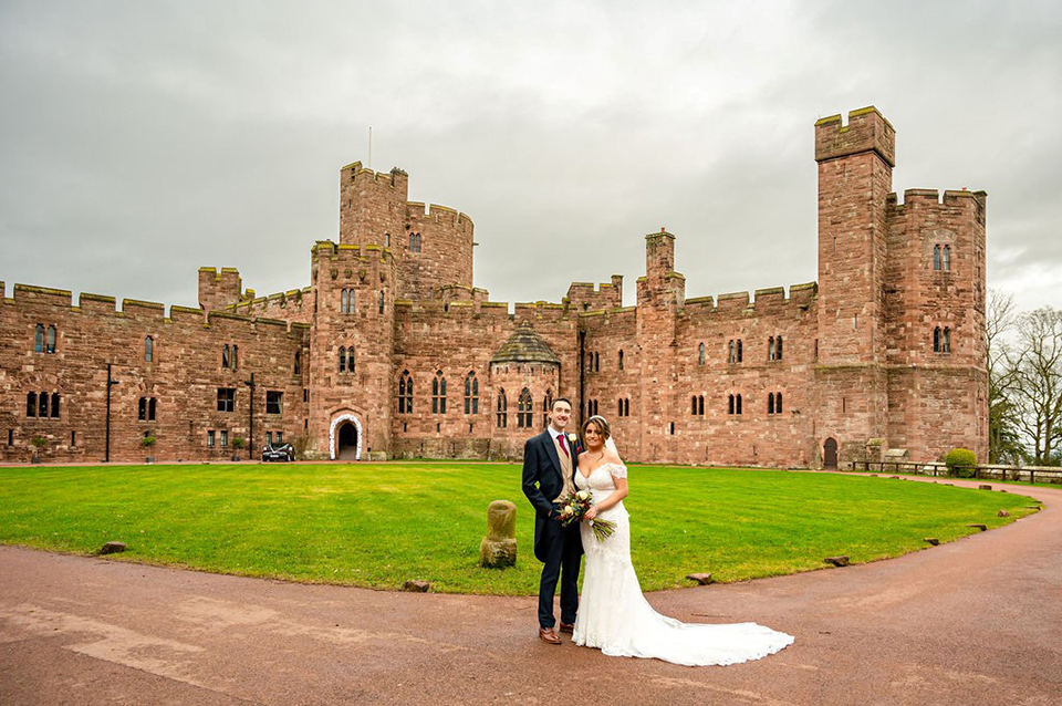A man wearing a suit and a woman in a white wedding dress posing outside the grounds of Peckforton Castle in Cheshire.